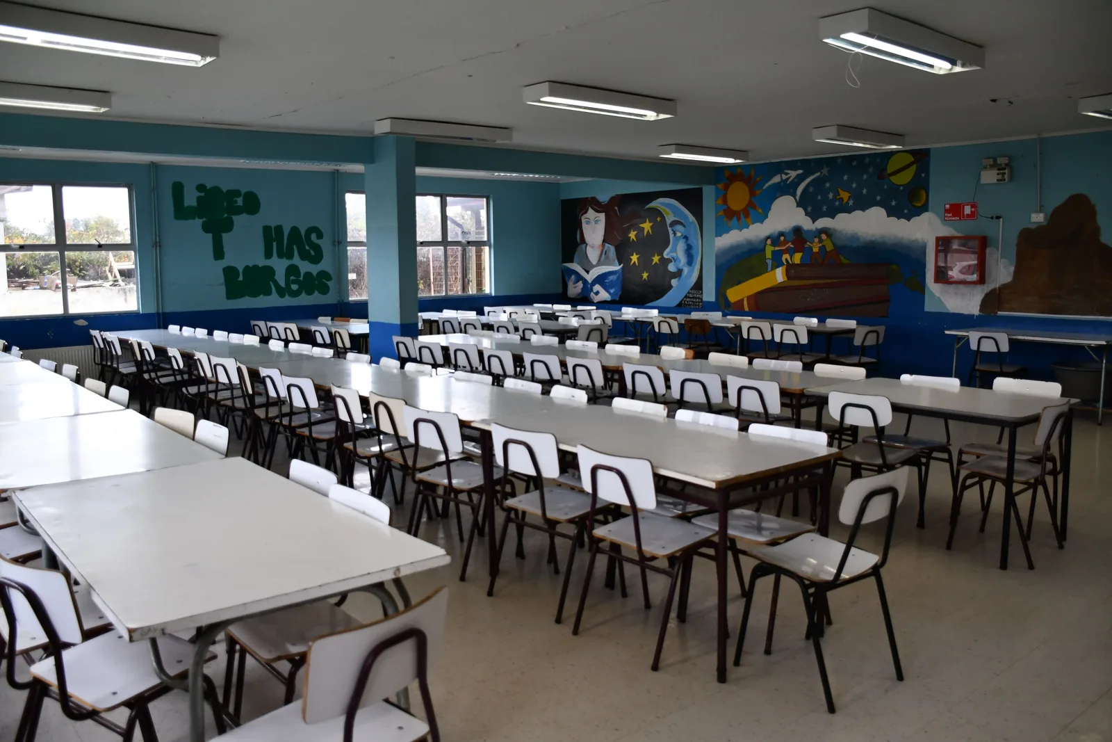 Comedor del liceo: mesas largas con sillas ordenadas, ventanales al patio y murales pintados en el muro del fondo.